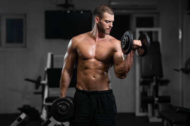 A man trains in the gym. Athletic man trains with dumbbells, pumping his biceps - Fotó, kép