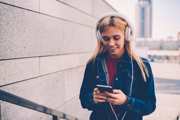 Smiling young woman in denim wear choosing music on internet website to download in playlist on phone.Positive student listening songs in headphones connected to cellular while chatting online - Fotoğraf, Görsel