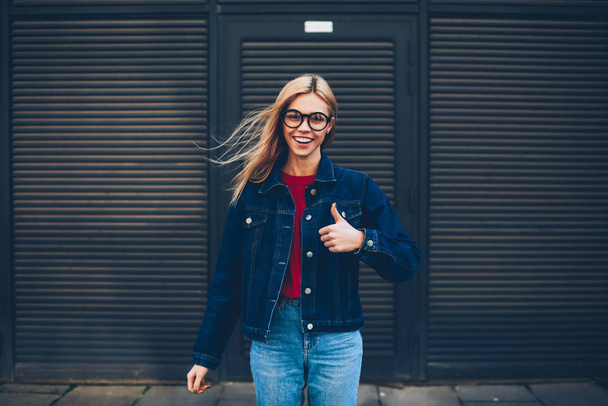 Portrait of cheerful blonde young woman in optical eyeglasses showing upper thumb up while smiling at camera standing on black promotional background.Positive hipster girl in denim jacket - Foto, Imagem