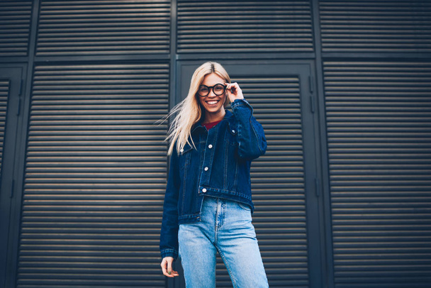 Below portrait of stylish positive young woman with blonde hair posing at camera.Cheerful hipster girl in optical eyeglasses dressed in denim clothes standing outdoors on black promotional background - Foto, Bild
