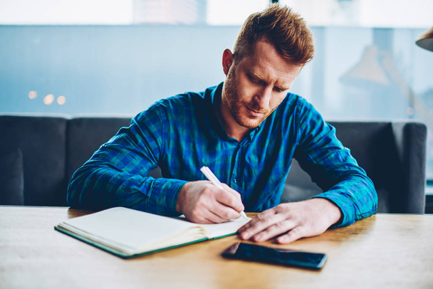 Skilled red haired student writing down homework in copybook studying at wooden table in coworking space.Pensive young man dressed in casual shirt noting checklist in notepad during free time - Photo, image