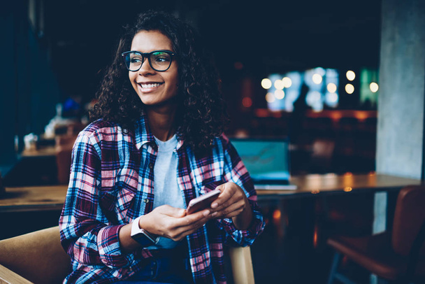 Belle afro-américaine hipster girl en lunettes souriant tout en envoyant un message SMS à un ami sur un appareil smartphone connecté à l'Internet 4G. Étudiante joyeuse à la peau foncée avec celluar dans les mains
 - Photo, image
