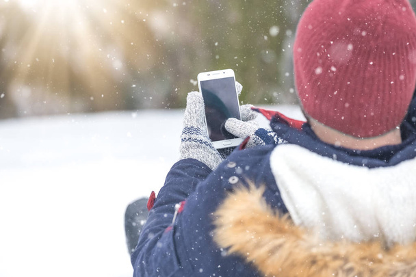 Rear view of man sitting in snow and holding blank screen smart phone. - Foto, Imagem