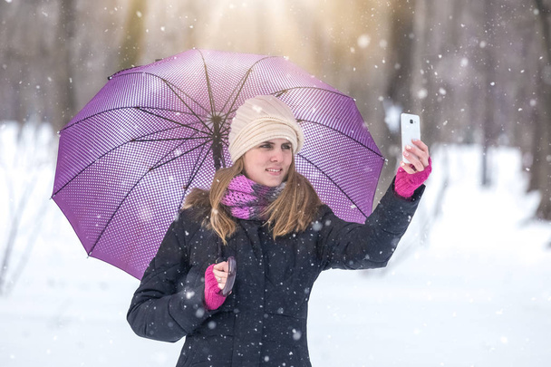 Woman with umbrella taking selfie with smartphone and enjoying winter season in park. - Zdjęcie, obraz