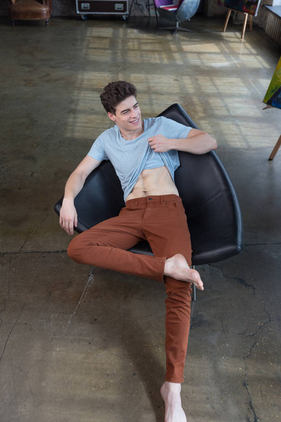 Handsome dark haired male model in blue t-shirt and brown pants sitting in a chair in a modern apartment - Foto, afbeelding