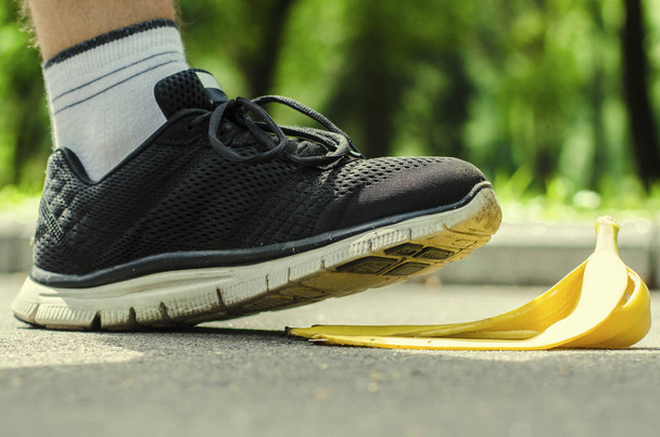 Il piede degli uomini in scarpe da ginnastica nere sature scivola su una buccia gialla da banana sullo sfondo di parco estivo
. - Foto, immagini