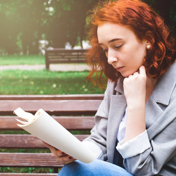 A girl is reading a book on a park bench. The girl has red hair and blue jeans - Foto, Imagem