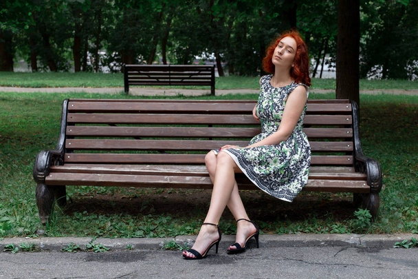 Roodharige meisje in een jurk zit op een bankje in een oud park. Concept - recreatie in stadsparken. in een oud park. Concept - recreatie in stadsparken. - Foto, afbeelding