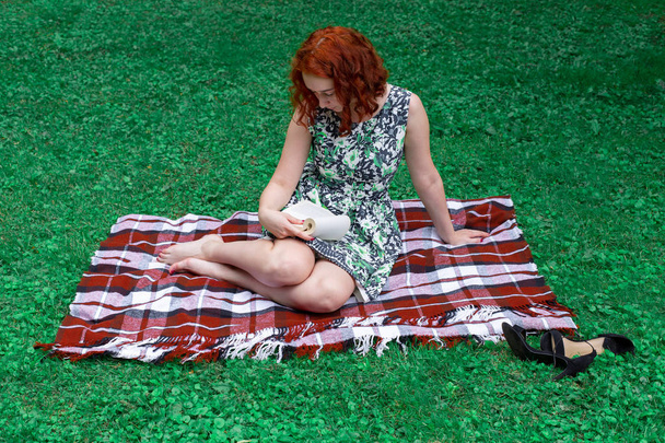 Red-haired girl in a city park on the grass reading a book - a literary novel. The girl has red hair. She sits on a woolen blanket. - Fotoğraf, Görsel