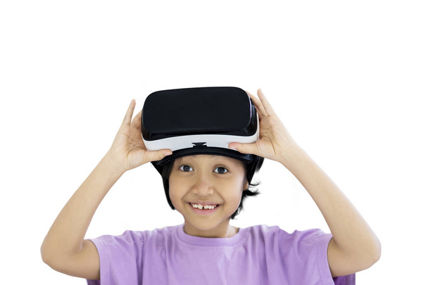 Image of adorable schoolgirl smiling at the camera while holding a virtual reality goggles, isolated on white background - Foto, Imagem