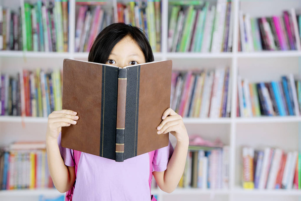 Picture of a cute schoolgirl looking at the camera and covering her face with a book while standing in the library - Foto, immagini