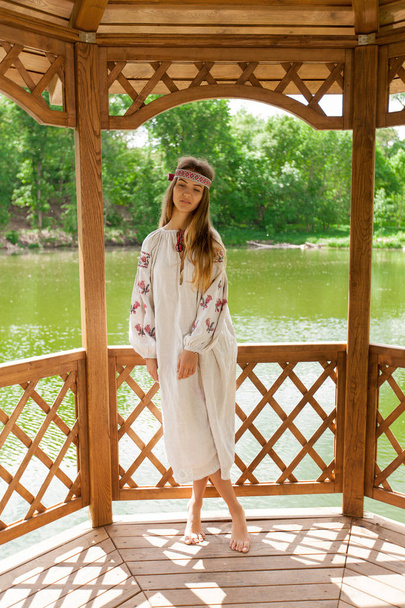 Beautiful Ukrainian woman dressed in embroidery in a wooden summer altanka on a lake - Photo, Image