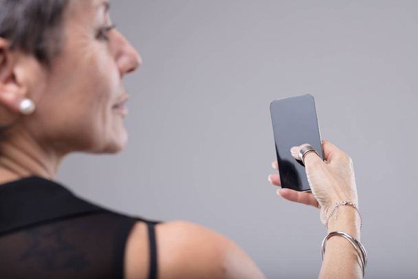 Stílusos nő, aki egy mobil a kezében egy több mint a váll érdekében az üres képernyő, ahogy fordul a fejét izolált szürke - Fotó, kép