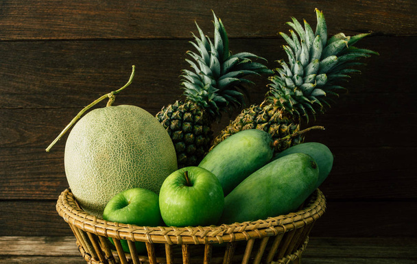 Fruta fresca, melón, mango, piña, manzana en canasta con luz natural y fondo de madera oscura
. - Foto, Imagen