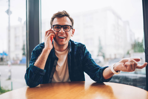 Young handsome emotional smart male student in trendy eyeglasses having fun while communicating with friends via smartphone.Portrait of cheerful man talking on mobile phone and looking at camera - Photo, image