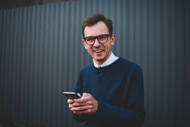 Portrait of cheerful male person in trendy eyeglasses having fun while sending messages to friends using telephone.Positive hipster guy laughing during summer walk at urban setting with gadget - Zdjęcie, obraz