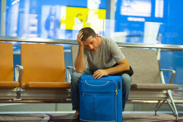 lifestyle portrait in airport of young attractive and tired tourist man with suitcase sitting worried and stressed at boarding gate waiting for cancelled or delayed flight ready for holidays travel - Foto, Imagen