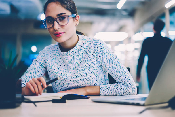 Portrait of skilled female employee in spectacles creating report sitting at desktop with modern laptop computer,pensive woman looking at camera analyzing information for startup at working  place - 写真・画像