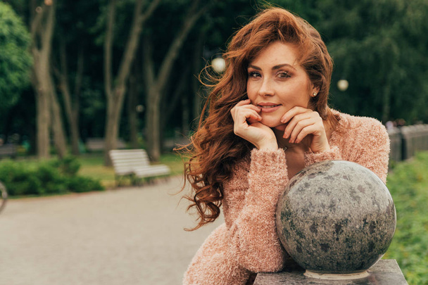beautiful, red-haired girl wearing warm clothes, leaning her elbows on a granite ball in the park, smiling at the camera - 写真・画像