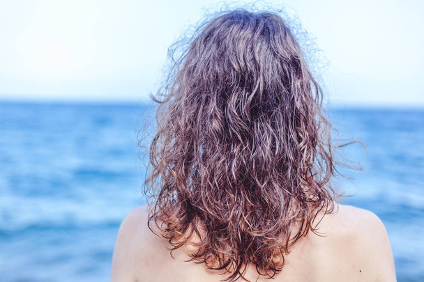 Curly haired brown-haired girl standing on the beach with her back, hair care at sea concept - Photo, Image