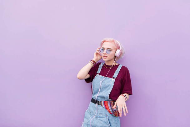 Стильная девушка-хиппи с розовыми волосами стоит на фоне фиолетовой стены и слушает музыку в наушниках. Музыкальная концепция
. - Фото, изображение