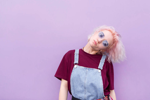 Portrait d'une fille à la mode en vêtements élégants, cheveux roses et lunettes de soleil qui repose sur le fond d'un mur violet. Élégante fille heureuse posant sur un fond violet
. - Photo, image