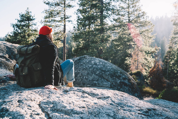 Back view of male traveler sitting on rock recreating during hiking tour enjoying wanderlust journey, hipster guy with rucksack looking at beautiful scenery of landscape exploring nature on weekend - 写真・画像