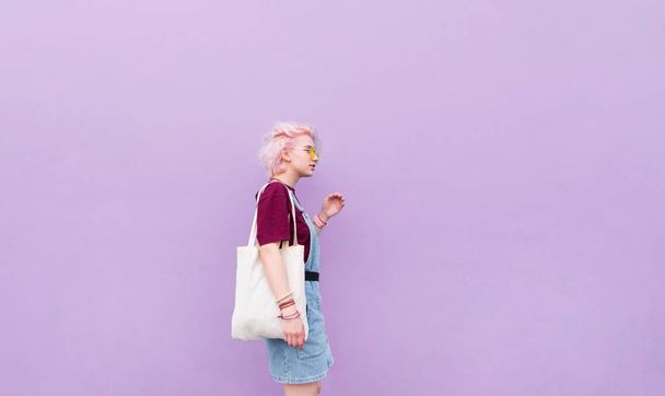 Stylish girl walking around the blue wall with a shopping bag. Girl with pink hair, glasses and shopper on the background of a purple wall - Photo, image
