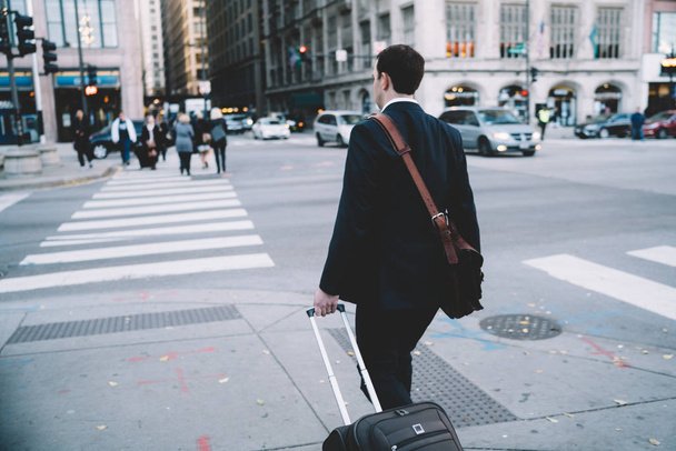 Back view of male entrepreneur in formal wear walking on avenue nera crossroads in downtown carrying suitcase, businessman strolling with luggage on urban settings of megalopolis during travel - Foto, imagen