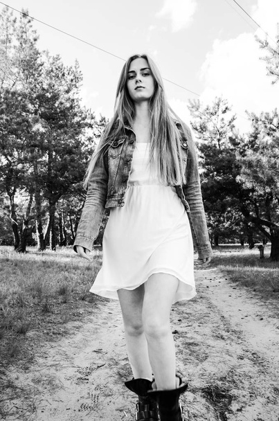 Maaseudun tyttö kävelee puulla hiekkatiellä. Tyttö pukeutunut valkoinen mekko ja sininen farkut takki. Nuori nainen liikkuu ja katsoo kameraa. Muotokuva. Keskikokoinen laukaus. Ulkona. Mustavalkoinen
. - Valokuva, kuva