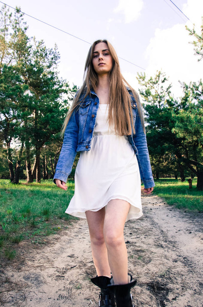 Maaseudun tyttö kävelee puulla hiekkatiellä. Tyttö pukeutunut valkoinen mekko ja sininen farkut takki. Nuori nainen liikkuu ja katselee kameraa
 - Valokuva, kuva