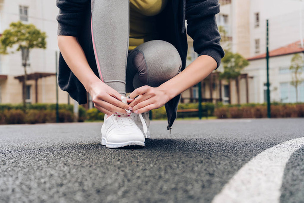 молодая девушка в серых спортивных брюках завязывает шнурки на кроссовках, готовится к тренировкам
 - Фото, изображение