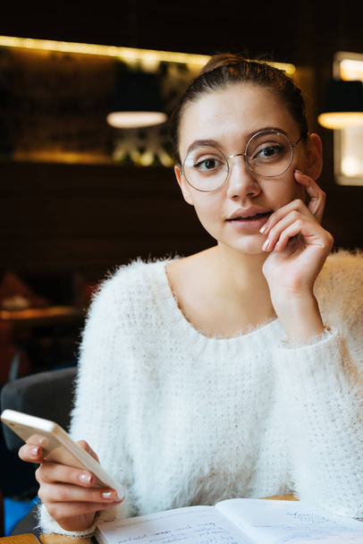 charming beautiful girl with glasses and a sweater resting in a cafe after work, holding a smartphone - Photo, image