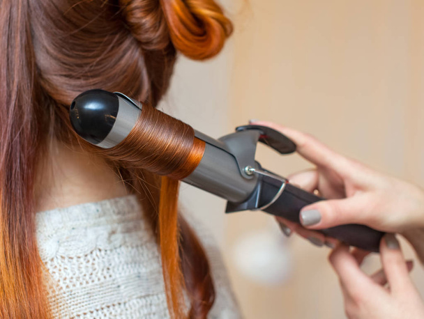 Κομμωτήριο κάνει hairstyle κορίτσι με μακριά κόκκινα μαλλιά σε ένα σαλόνι ομορφιάς. Δημιουργία μπούκλες με σίδερα κέρλινγκ. Επαγγελματική περιποίηση μαλλιών. - Φωτογραφία, εικόνα