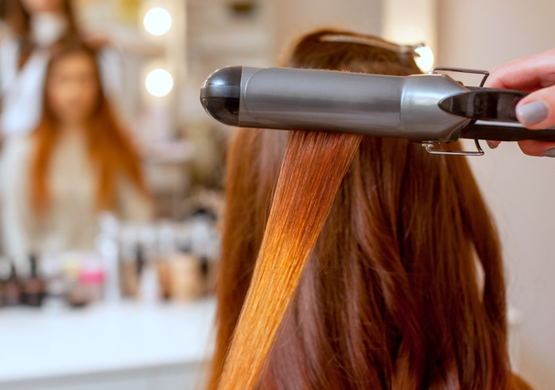 Kampaaja tekee kampauksen tyttö pitkät punaiset hiukset kauneushoitola. Luo kiharat curling silitysraudat. Ammattimainen hiustenhoito. - Valokuva, kuva