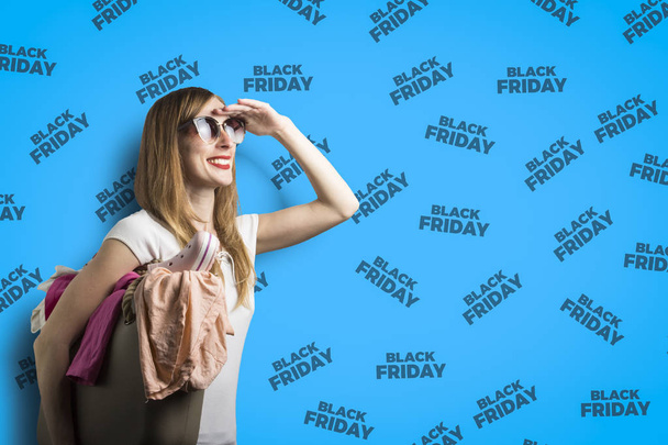 Una joven en gafas con una bolsa llena de ropa mira a la distancia en anticipación de Black Friday sobre un fondo azul con el texto Black Friday
. - Foto, Imagen
