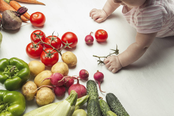 Little baby looks at fresh organic vegetables on a light wooden background. Care for health. Choice of the first food. Healthy baby food. Flat lay, top view. - Zdjęcie, obraz