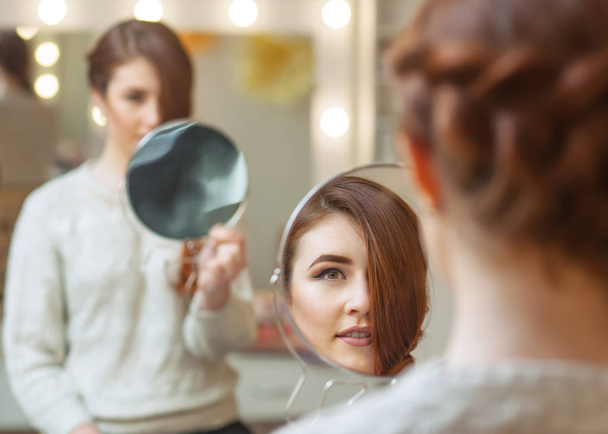 Retrato de una hermosa chica pelirroja con el pelo largo, que se mira en el espejo en el salón de belleza. Cuidado profesional del cabello
 - Foto, Imagen
