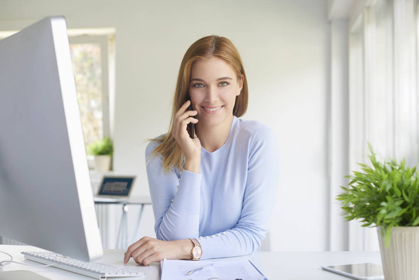 Beautiful young businesswoman talking with someone on her mobile phone while sitting in front of computer.  - Foto, Bild