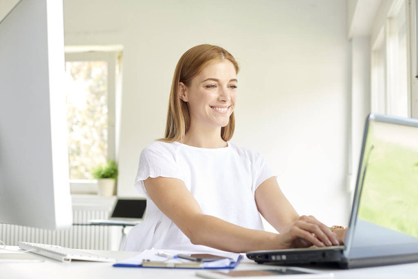 Shot of smiling young businesswoman using her laptop while sitting at office desk and working online.  - Foto, Bild