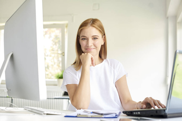 Tiro de sorridente jovem empresária usando seu laptop enquanto está sentado na mesa de escritório e trabalhando online.  - Foto, Imagem