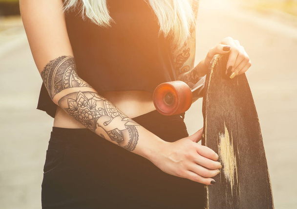 Ένα hipster όμορφη, σέξι ξανθιά με μπλε μαλλιά τατουάζ στέκεται με ένα longboard στο δρόμο. - Φωτογραφία, εικόνα