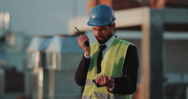 Young man supervisor at construction site speaking from radio and analyzing the construction plan from tablet. - Video, Çekim