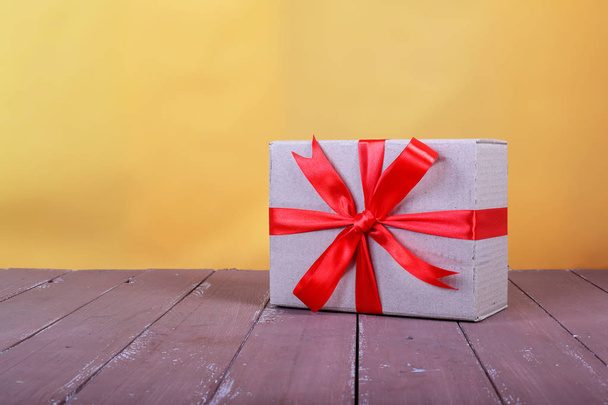 Servicio de envío de correos y embalaje: pequeño paquete atado por un lazo rojo sobre un fondo de madera marrón y pared amarilla
. - Foto, Imagen