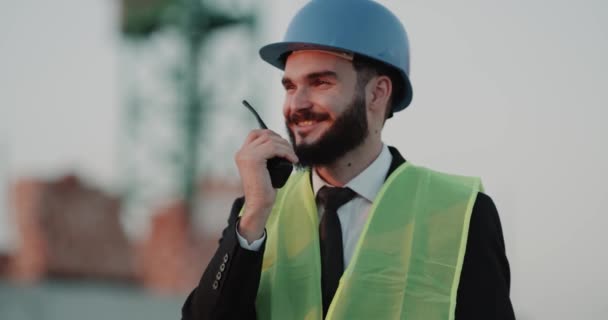 Young engineer at construction site wearing a safety helmet and speaking with his workers using a radio - Кадры, видео