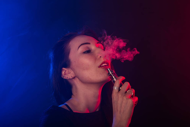 Portrait of young woman in neon red and blue smoke with vape or e-cigarettes - Photo, Image