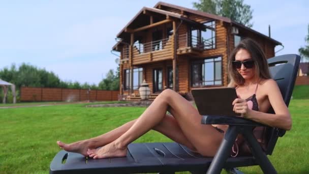 A beautiful sexy woman lying on a sun lounger in the backyard of her mansion is doing online shopping with a tablet computer - Footage, Video