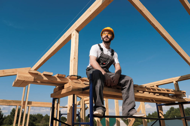 The man builder sits on the edge of the roof of the frame house, in a yellow helmet and gray overalls. The blue sky and clear sunny day. - Фото, изображение