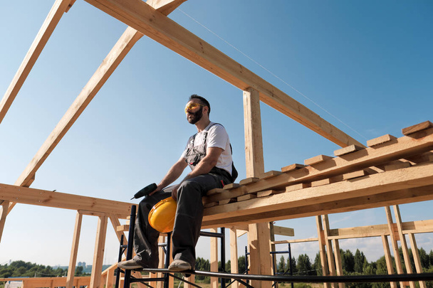 The man builder sits on the edge of the roof of the frame house, in a yellow helmet and gray overalls. The blue sky and clear sunny day. - Photo, image