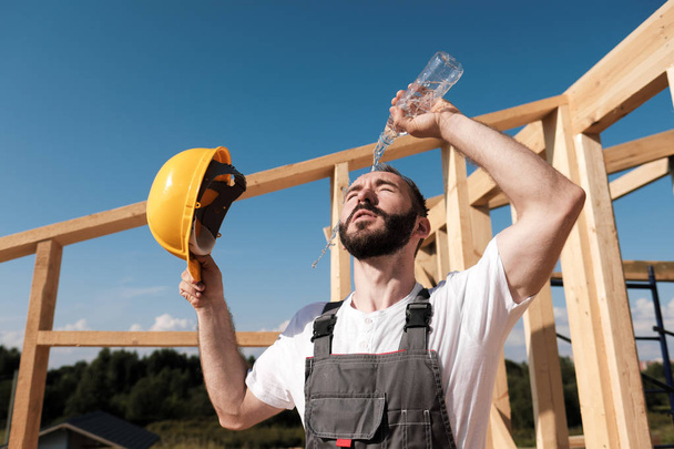 The man builder sits on the edge of the roof of the frame house, in a yellow helmet and gray overalls and a white T-shirt. He pours water on the head from the bottle. - Foto, Imagem